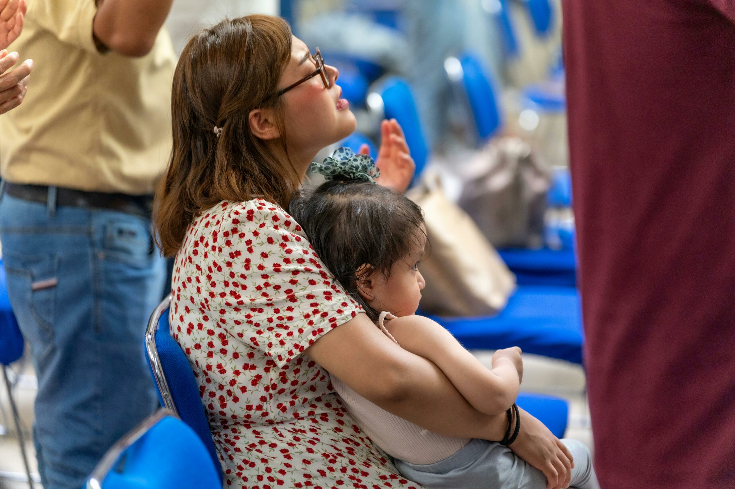 pexels-israwmx-24023650 image of a medium length haired person with a child in her lap, seated in a room with other chairs and people