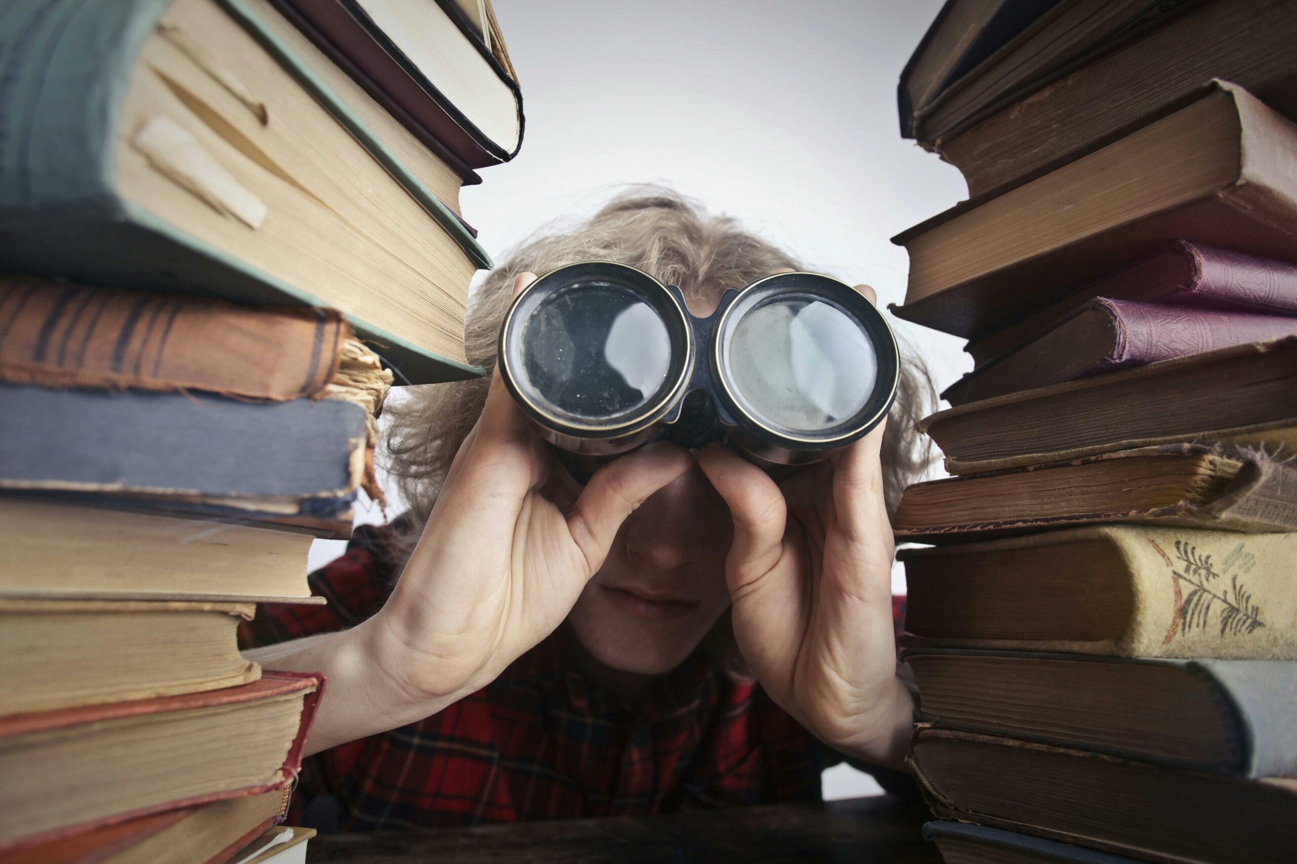 pexels-olly-3769697 a white person peering through binoculars at the viewer with piles of books on either side